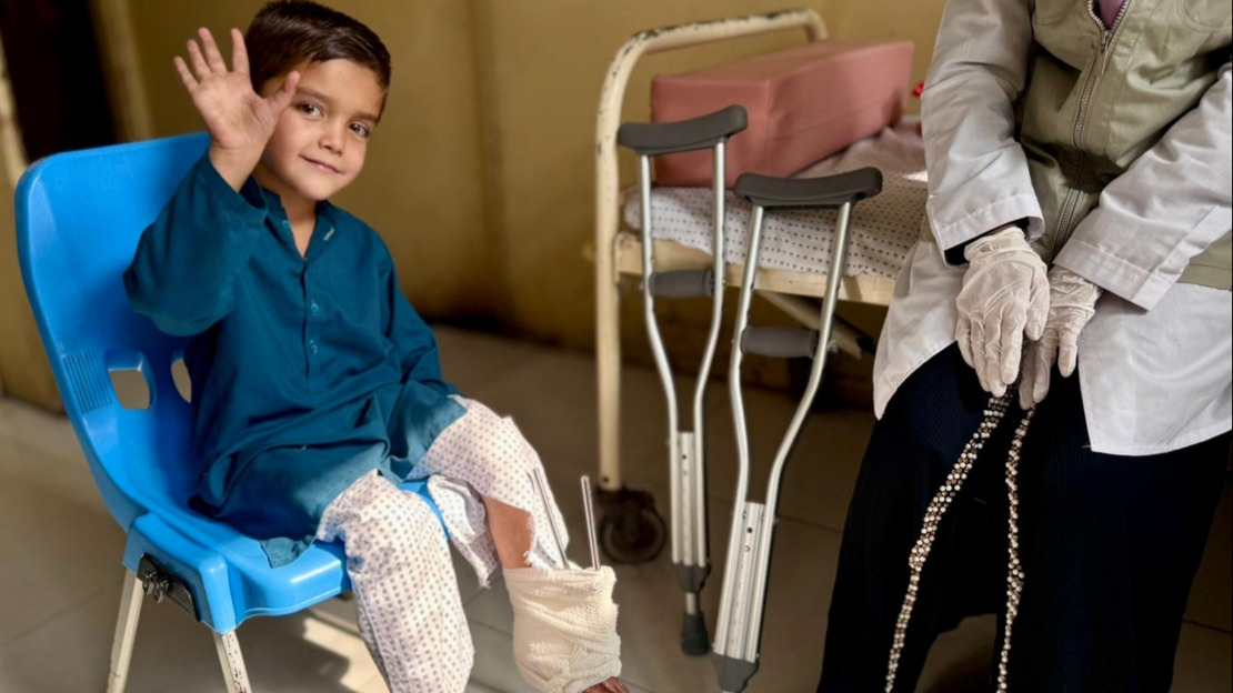 A little boy is sitting on a chair and waving at the camera. His foot is bandaged and crutches are placed next to him.