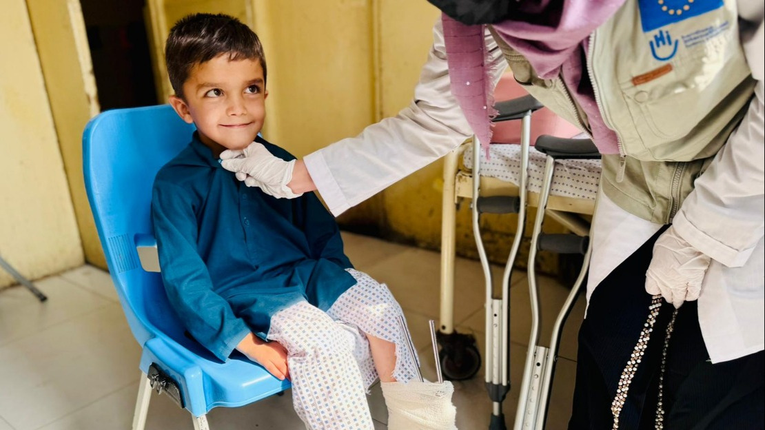 A smiling little boy is sitting on a chair and looking at a woman leaning over him who is wearing a HI/European Union jacket.