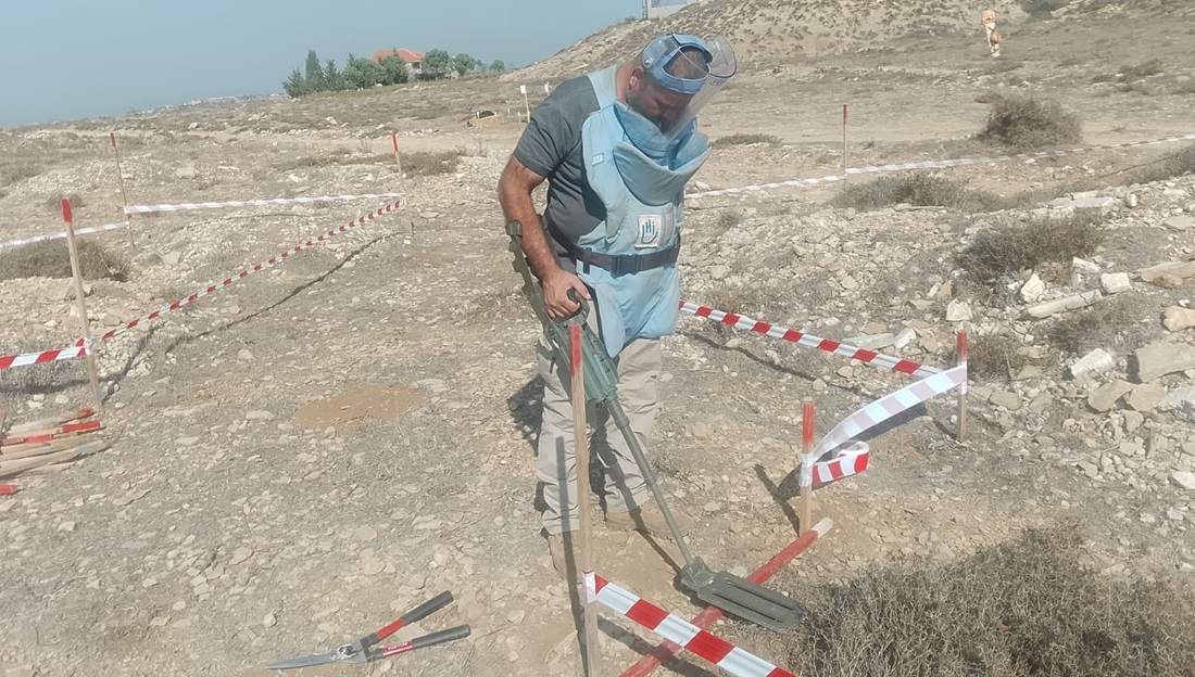 An HI deminer is clearing a plot of land identified as contaminated with explosive devices. He is standing and holding a detector.