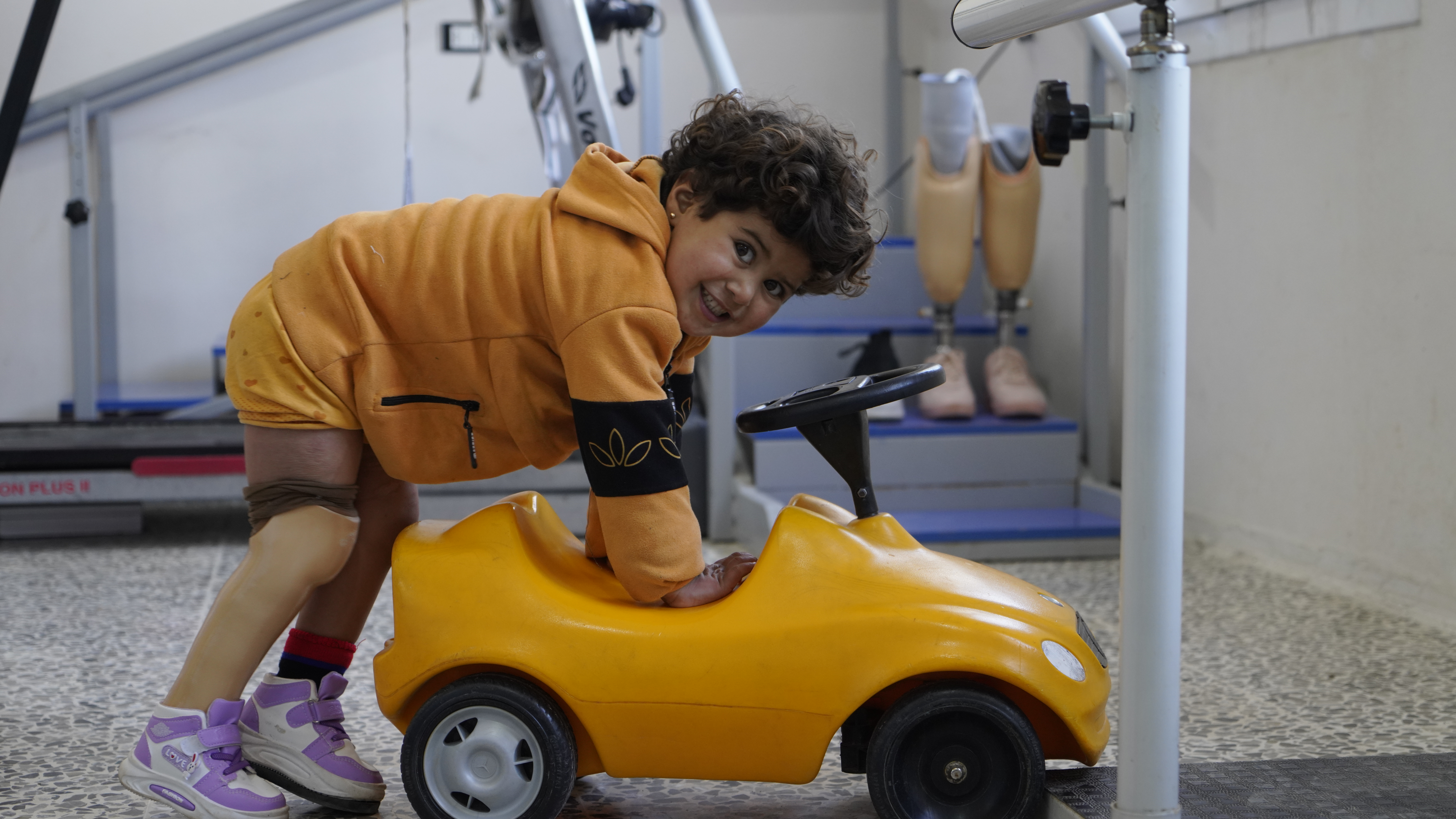 Noor during a rehabilitation session she jumps on a little yellow car smiling at the camera at Aqrabat hospital, HI’s partner in Northwest Syria. 