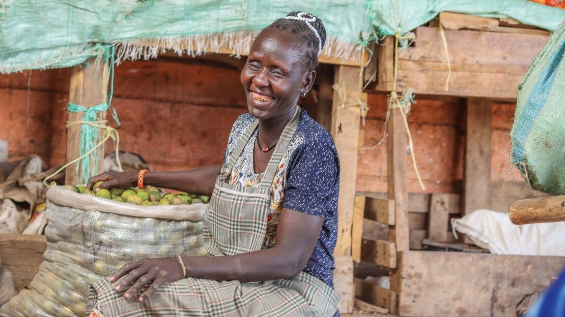 A woman sits laughing. Her right hand is resting on a large canvas bag containing fruit. Behind her are stalls of fruit and vegetables.