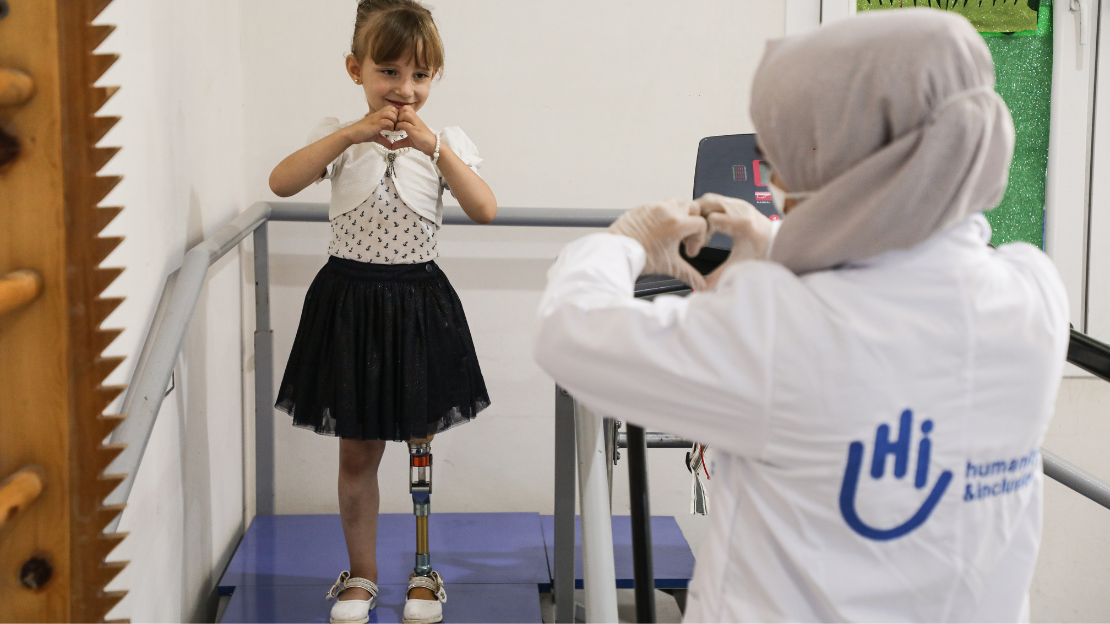 Enas has a prosthetic leg and stands on a platform, making a heart shape with her hands. She is facing a woman wearing a white lab coat and headscarf, who mirrors the gesture. The coat has the HI logo printed on the back. They are in a bright room with metal railings, suggesting a rehabilitation setting.