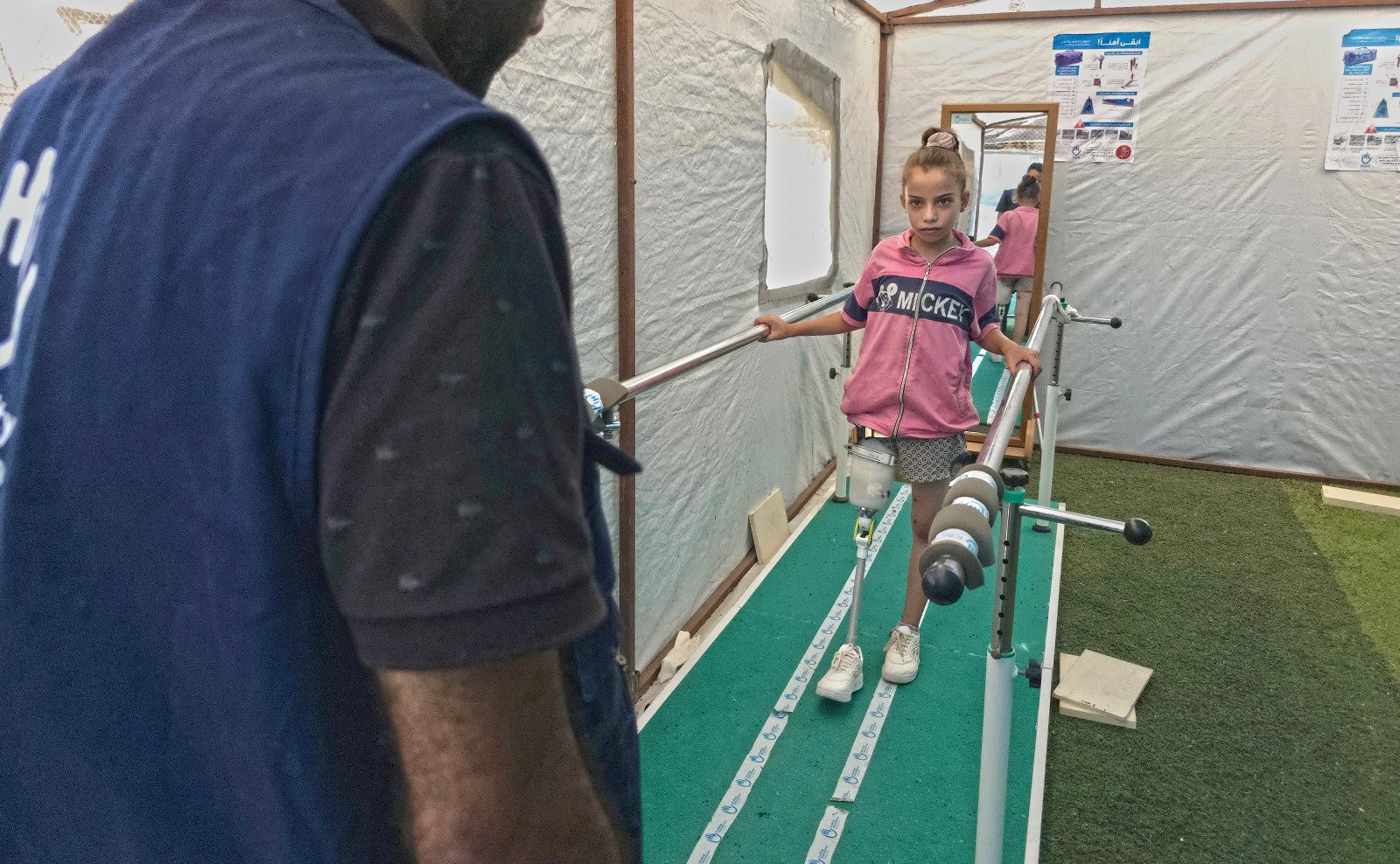 Malak stands in HI's rehabilitation centre in Gaza, supported by two bars. She is learning how to walk using a prosthetic leg.