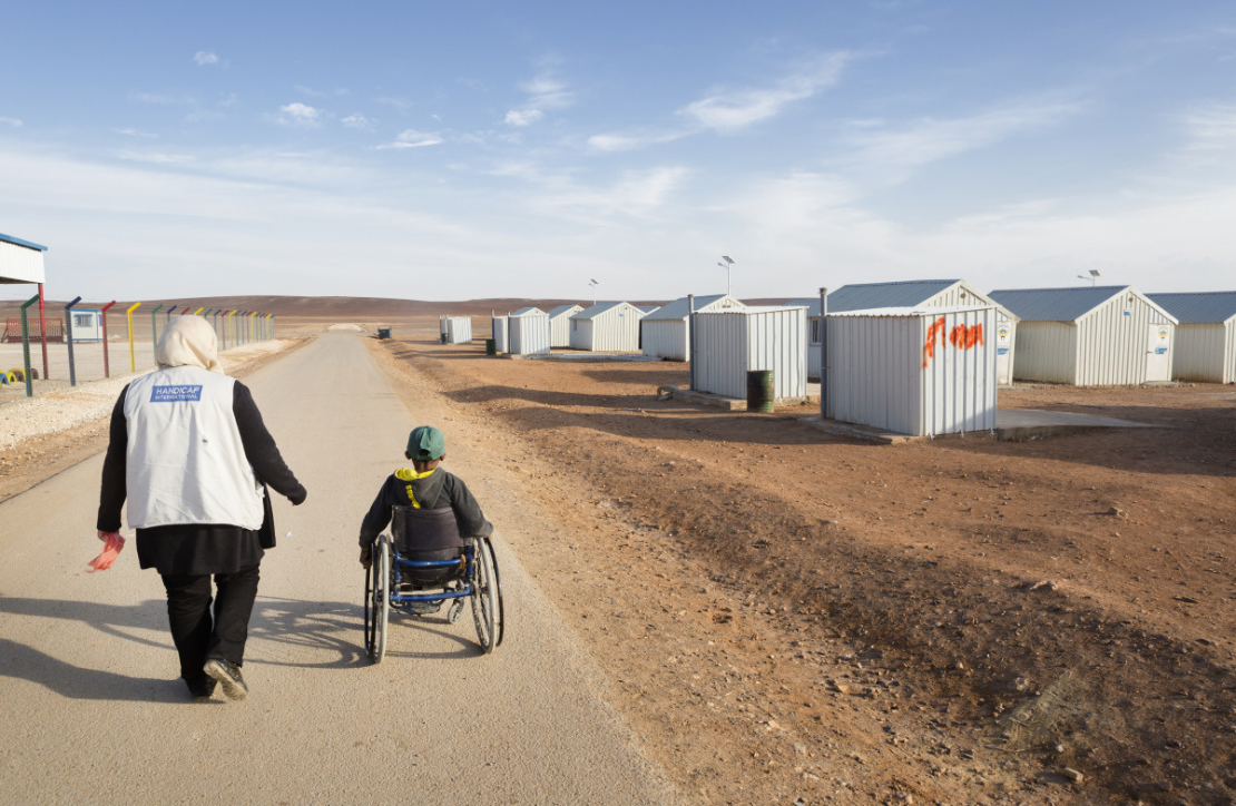 A member of HI staff walking alongside a Syrian boy in a wheelchair in Azraq camp, Jordan