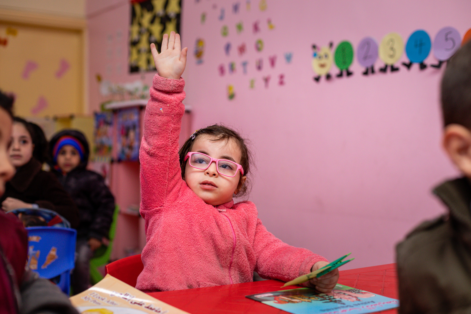 Sham, 5, with her right hand raised in her classroom. She wears spectacles and a pink jumper.
