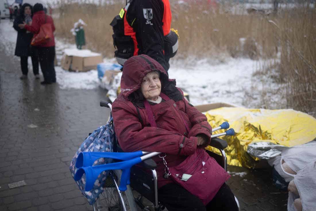 An 87 year-old woman in a wheelchair with a pair of crutches resting across her lap. There is snow on the ground. She is crossing the border from Ukraine to Poland.