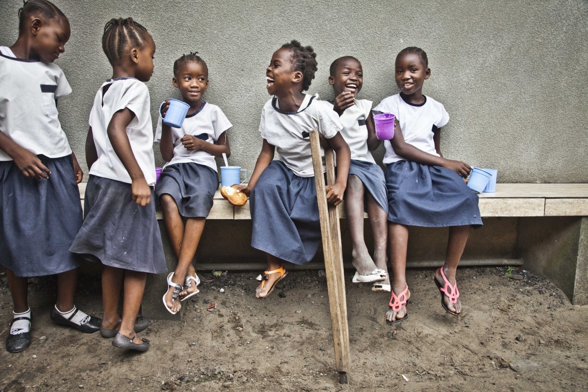 Grace, a young girl who is holding a wooden crutch, is sitting on a bench and smiling. She is surrounded by her friends.