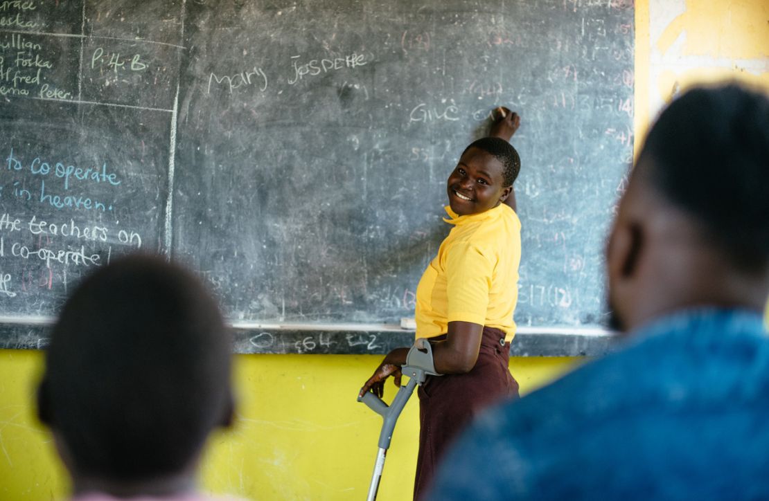 A girl using an elbow crutch writes on a chalkboard in a classroom, Uganda