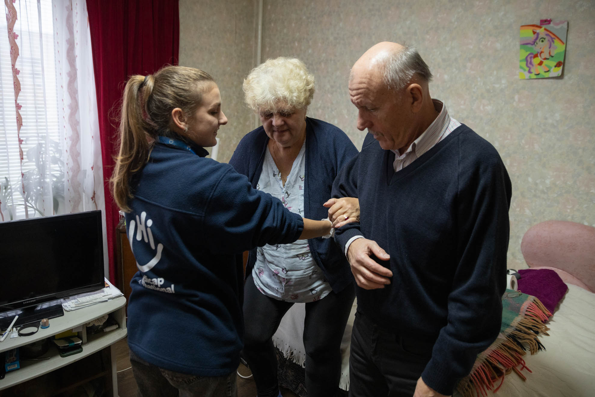 Antonina is holding her husband’s arm while a female HI staff is also supporting her. 