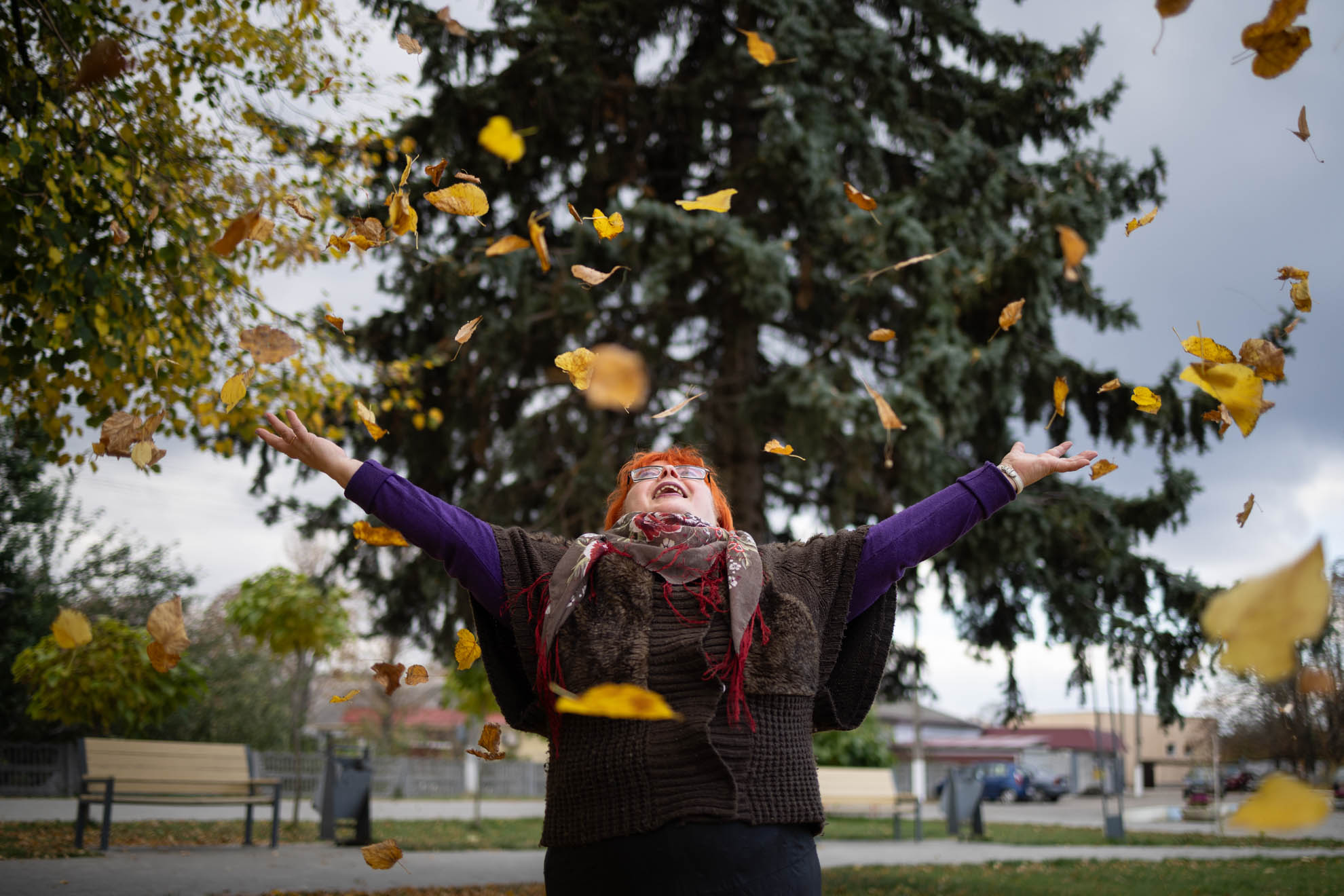 Olena is smiling in front a background of trees and greenery throwing leaves in the air. She has red hair, glasses and a colourful scarf. 