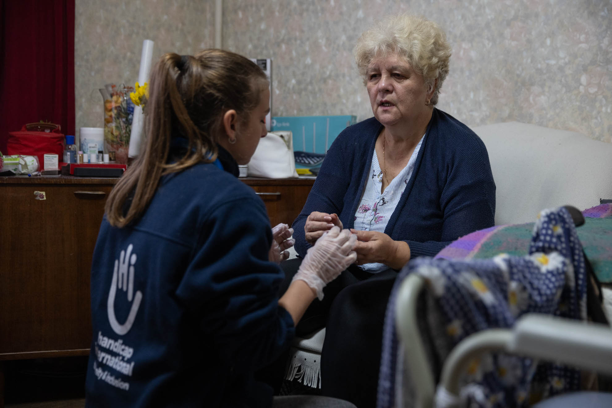 Antonina is sitting on an armchair and a female HI staff is holding her hand. 