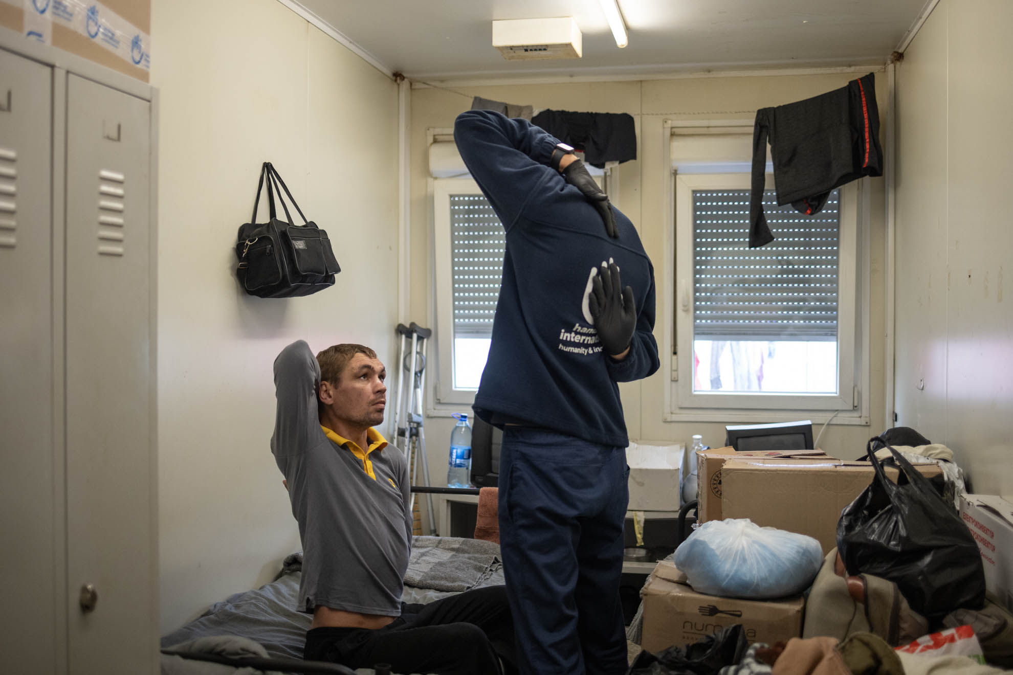 A male HI staff and Serhii are standing in front of his shelter, talking. HI’s staff has his hand on Serhii’s shoulder and is wearing a mask. 