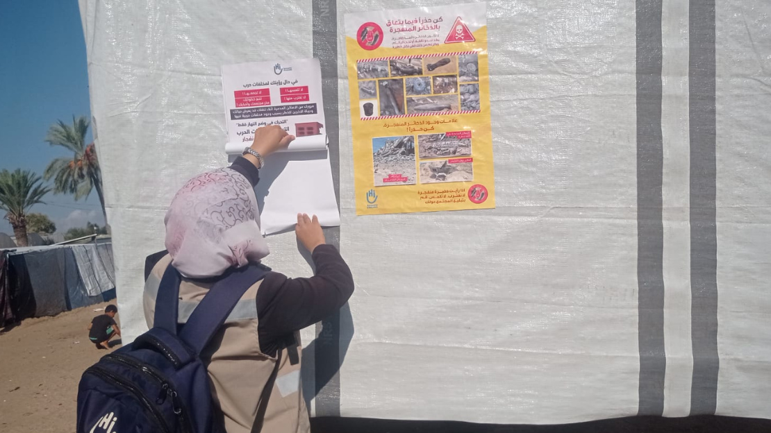 A HI staff woman, wearing a bag with the HI logo, is putting up a sign informing people about explosive remnants. She is in a refugee camp, sticking the sign on a wall. ; }}