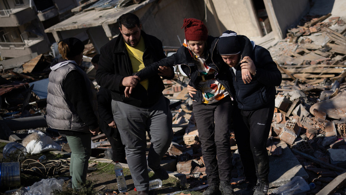 People walk in front of a collapsed building with a wounded person; }}