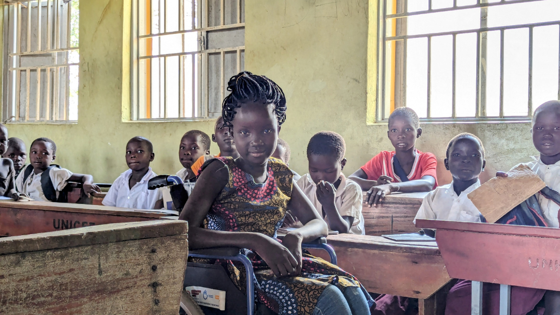 View of a classroom with students seated at desks. In the foreground, a smiling adult woman looks to her left at a young girl in a wheelchair.; }}