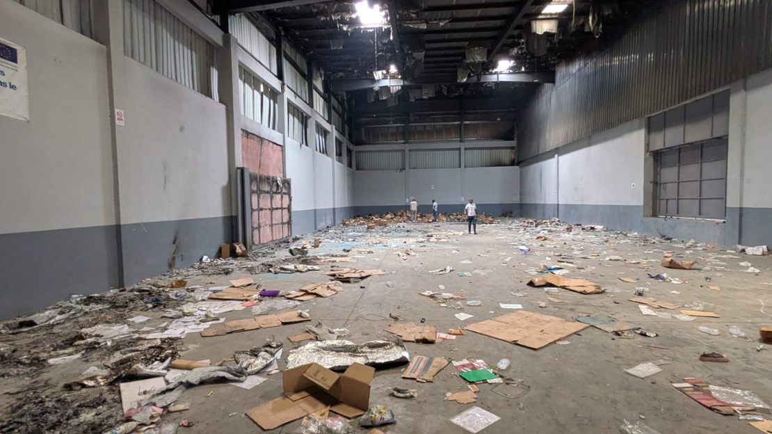 Inside a warehouse: the floor is littered with cardboard boxes, sacks and garbage of all kinds. Part of the roof is black with soot, and sheet metal panels are peeling off.; }}