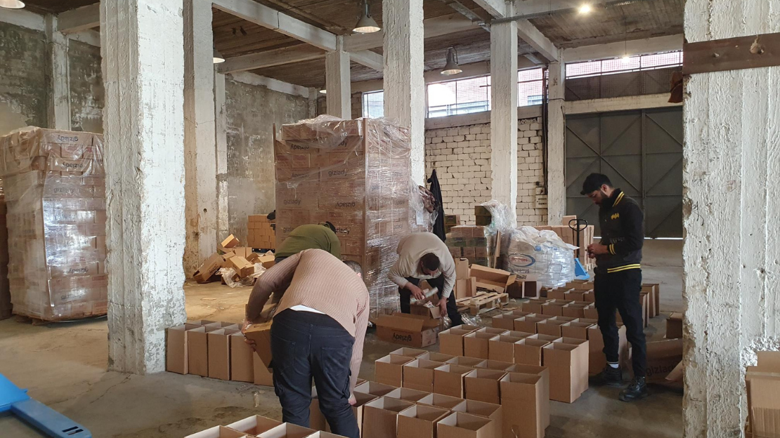 Three people are assembling cardboard boxes in a spacious, industrial-style warehouse with exposed concrete beams and a high ceiling. Stacks of packaged goods wrapped in plastic are visible in the background. The workers are focused, leaning over the boxes as they work. 
