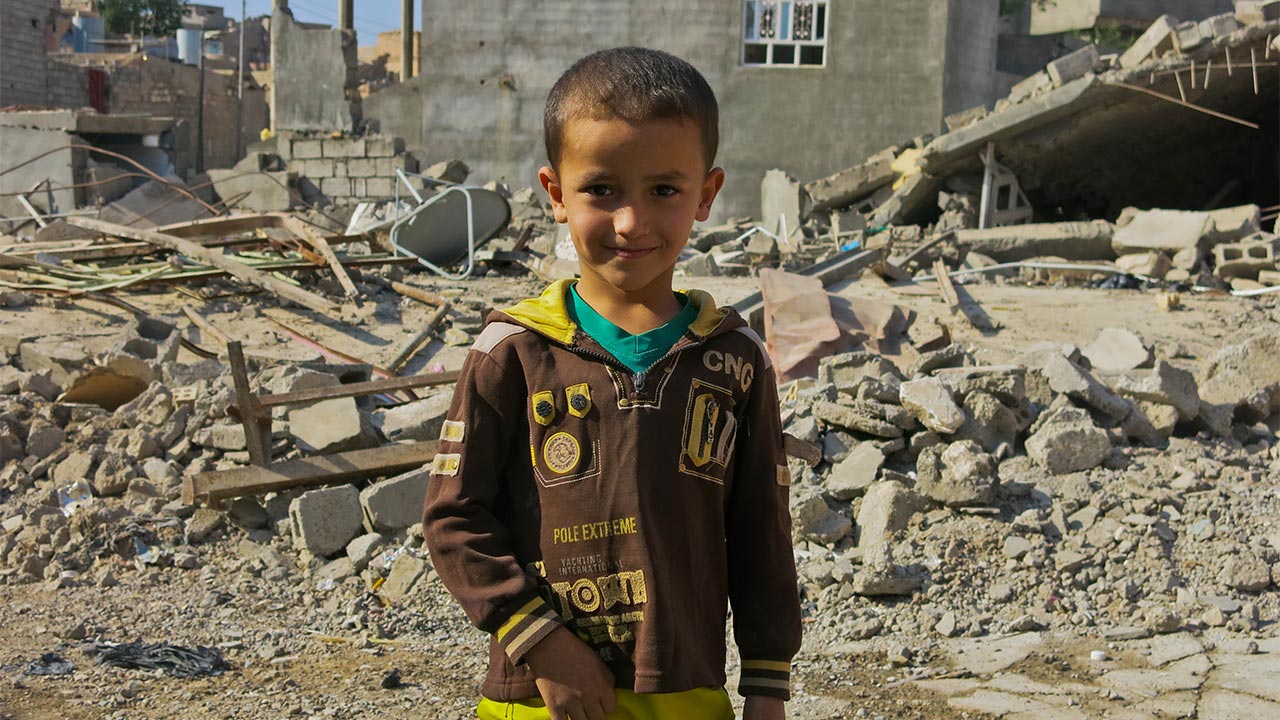 A child stands in front of damaged buildings in Jalawla, Iraq.; }}